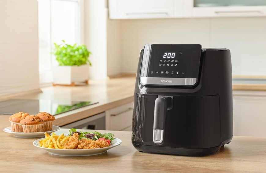 Fritéza teplovzdušná Sencor SFR 6550BK-EUE3 čierna Teplovzdušná fritéza Sencor SFR 6550BK-EUE3 je položená na svetlej kuchynskej linke. Vedľa nej sú položené stripsy s hranolkami, zeleninou a muffinmi.