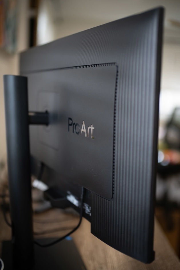Monitor Asus ProArt PA27UCGE čierny Na obrázku je zadná strana monitora Asus ProArt PA27UCGE 27" zachytená z bočného detailu. Monitor má čierny zadný kryt s jemným zvislým rebrovaním a uprostred plastový panel s logom ProArt. Vľavo je vidieť zvislý stojan a rameno, ktoré držia displej ku displeju. V spodnej časti zadnej strany sú pripojené kabely. matný dizajn.