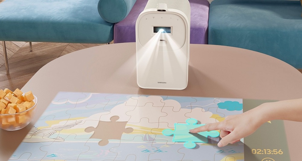 Laser TV Samsung SP-LPF5DSA Projektor The Premiere 5 v stojane stojí na stole a premieta naň puzzle, ľudská ruka posúva dielik puzzle