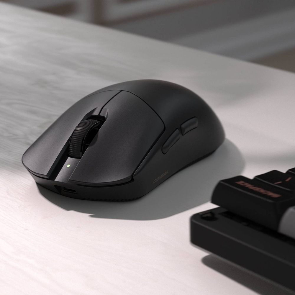 Myš Cougar Revenger PRO 4K čierna Na obrázku je čierna bezdrôtová herná myš Cougar Revenger PRO 4K položená na svetlom stole vedľa klávesnice. Myš má matný povrch, výrazne klenutý ergonomický tvar, vrúbkované rolovacie koliesko a malú zelenú stavovú kontrolku. Na ľavom boku sú vidieť dve bočné tlačidlá pre palec. V prednej časti je otvor na pripojenie kábla na nabíjanie alebo káblové použitie.