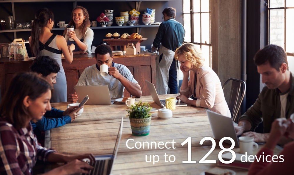 Router D-Link 5G NR AX3000 Wi-Fi 6 biely Útulná kaviareň, kde ľudia pracujú na notebookoch pri drevených stoloch. Atmosféra je produktívna a uvoľnená. Na spodnej strane obrázku vidíme text v znení: „Pripojte až 128 zariadení.“