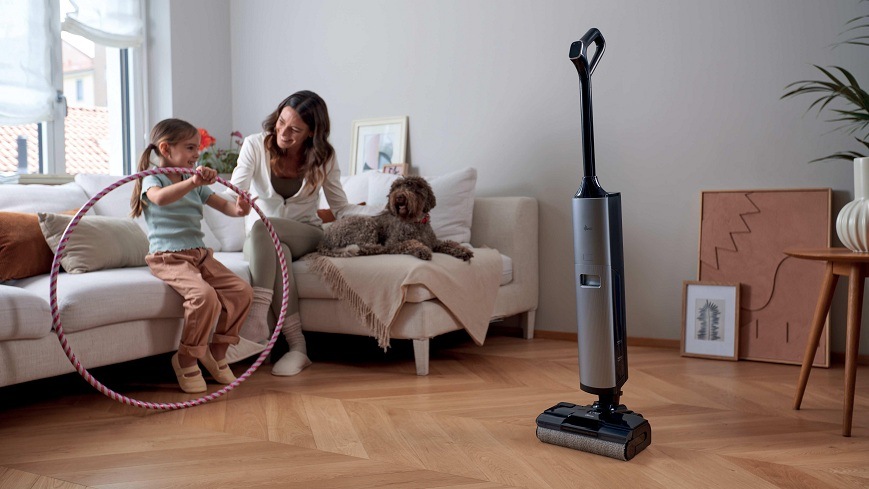 vysávač Hoover HW600 011 stojí uprostred miestnosti, zatiaľ čo žena s dcérou a psom sedí spolu na gauči