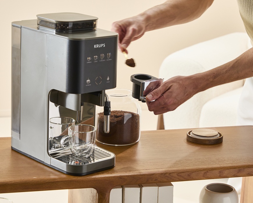 Automatický kávovar Krups Coffee Crush Experience SA403BE0 sivý/hnedý Žena má v ruke lyžicu a na ňu má mletú kávu. Tú dáva do zásobníka v automatickom kávovare Krups SA403BE0 Coffee Crush Experience.