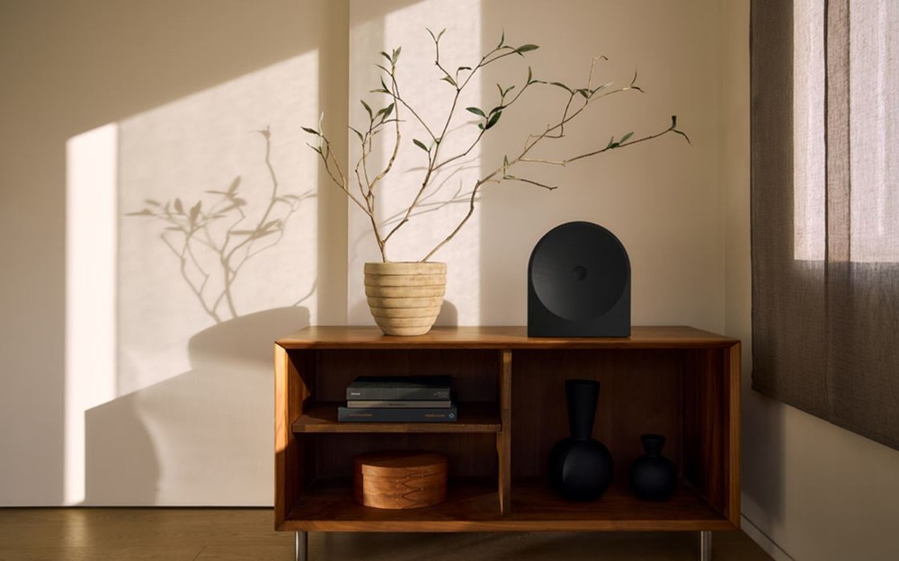 Reproduktor Samsung Music Studio 5 LS50H (1 ks) čierny Minimalistický interiér s čiernym reproduktorom Samsung HW-LS50H Studio 5 na drevenej skrinke vedľa kvetináča s vetvami, osvetlený teplým slnečným svetlom vrhajúcim tiene na stenu.