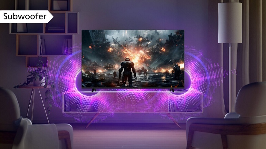 Televízia JVC umiestnená v modernej obývacej izbe, na ktorej obrazovke je zobrazená scéna z akčnej videohry s futuristickým vojakom. Z dolnej časti televízora sú fialovými vlnami zvýraznené zvukové efekty, ktoré naznačujú silný basový výkon vstavaného subwoofera. V ľavom hornom rohu je popis "Subwoofer"