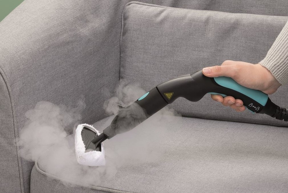 Čistenie šedého gauča pomocou parného čističa Ariete ART 4147/BL XVapor s nástavcom a návlekom na čalúnenie.