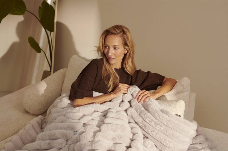 Vyhrievacia deka Duux DXOB02 Yentl Cuddle 200 x 200 cm sivá Žena pololeží v širokom kresle v teplom večernom osvetlení, zababušená do mäkkej béžovej vyhrievacej deky Duux Yentl, v pozadí vysoká izbová rastlina a pokojná domáca atmosféra.
