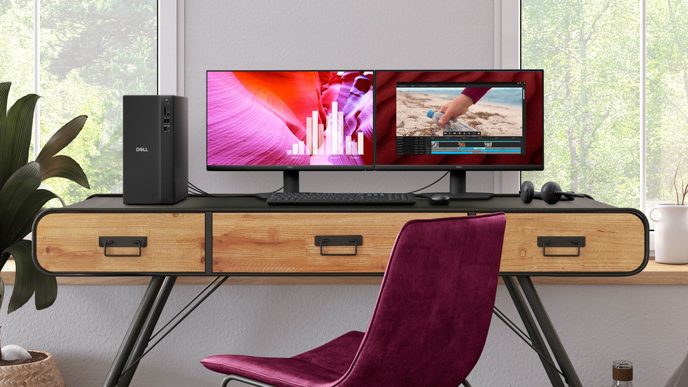 Stolný počítač Dell Pro Tower Essential QVT1260 čierny Moderný pracovný priestor s dreveným stolom, dvoma monitormi, stolným počítačom a slúchadlami na boku. Pred stolom stojí ružová stolička a z okien je vidieť zeleň.