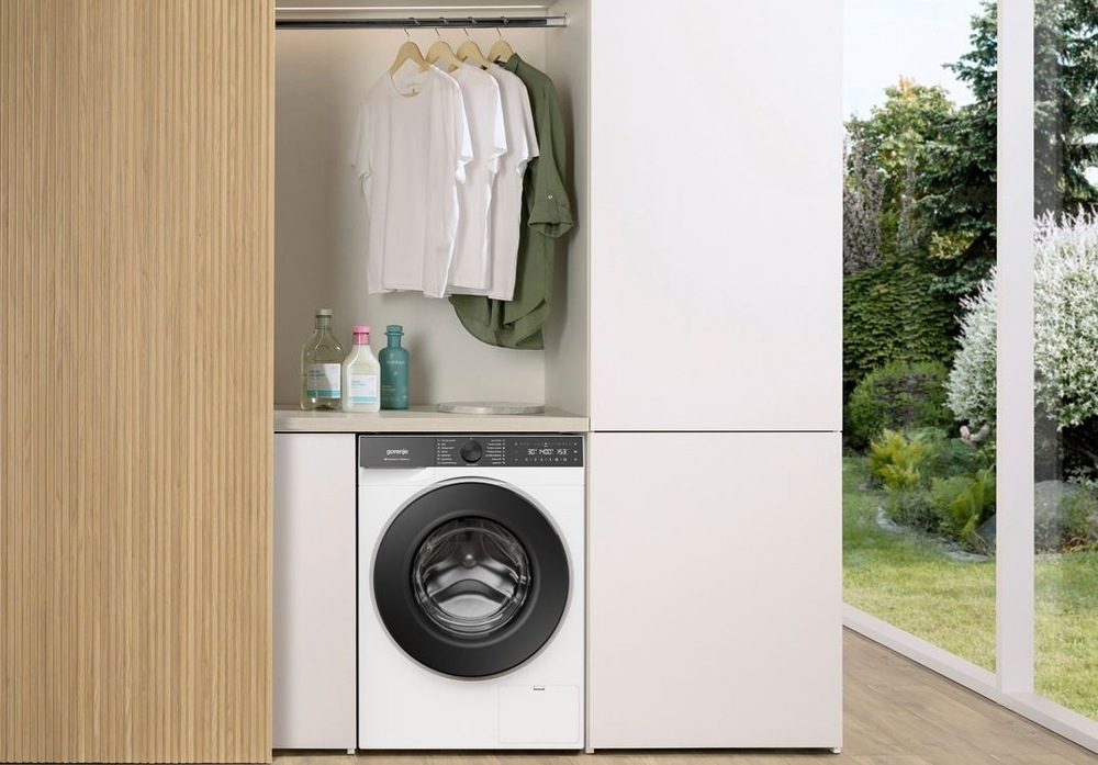 Práčka Gorenje G600 WG614A4P1 FastWashPro biela práčka Gorenje WG614A4P1 FastWashPro zabudovaná v drevenej skrinovej stene. Nad ňou visi na ramienku tričká. Vedľa sú presklené dvere do záhrady