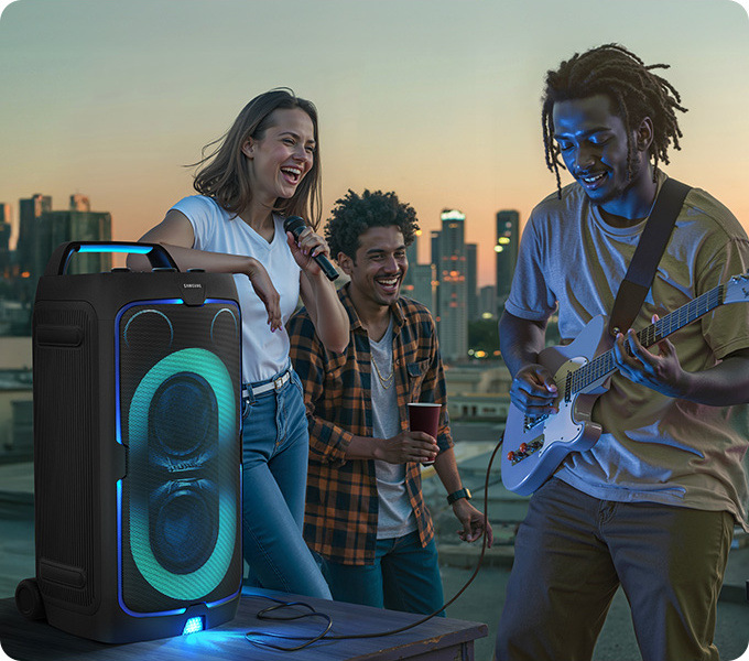 Párty reproduktor Samsung Sound Tower MX-ST50F čierny Do reproduktora Samsung MX-ST50F je zapojený mikroforn a elektrická gitara, traja ľudia stoja pri reproduktore a spoločne hrajú a spievajú