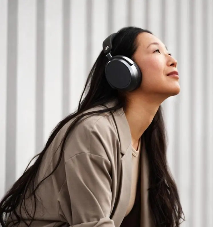 Žena sa zamyslene pozerá pred seba, na ušiach má slúchadlá Sennheiser MOMENTUM 4″ width=