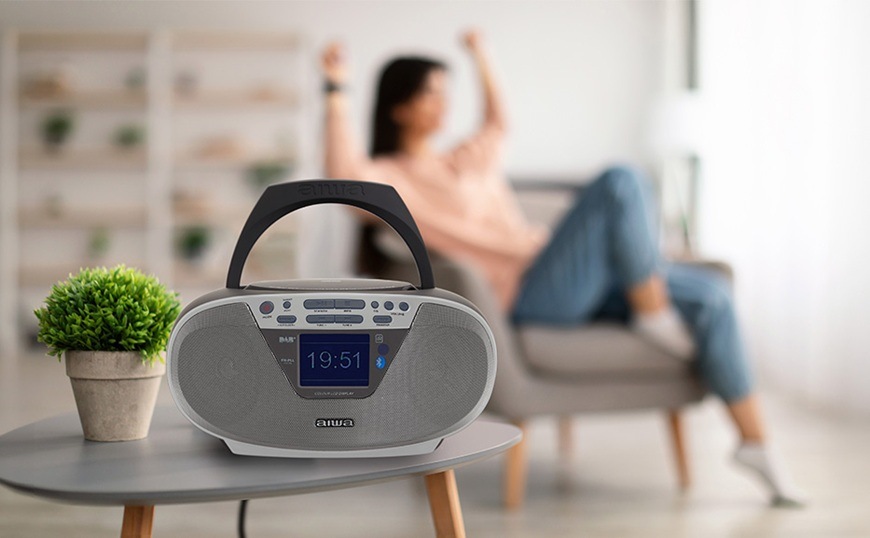 Rádiomagnetofón s DAB+/CD AIWA BBTU-500DAB strieborný Rádioprijímač DAB+/CD Aiwa BBTU-500DAB/SL je položený na konferenčnom stole. Žena v pozadí sedí v kresle a počúva svoju obľúbenú hudbu.