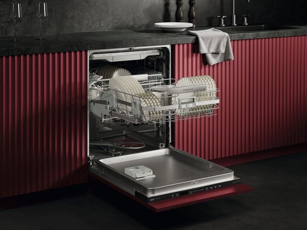 Vstavaná umývačka AEG NG82IA50TK séria 8000 CleaningPerformance otvorená. Spodný kôš je vďaka závesov ComfortLift v úrovni horného koša a je plne naložený taniermi. Umývačka je vsadená do kuchynskej linky s červeným dekórom.