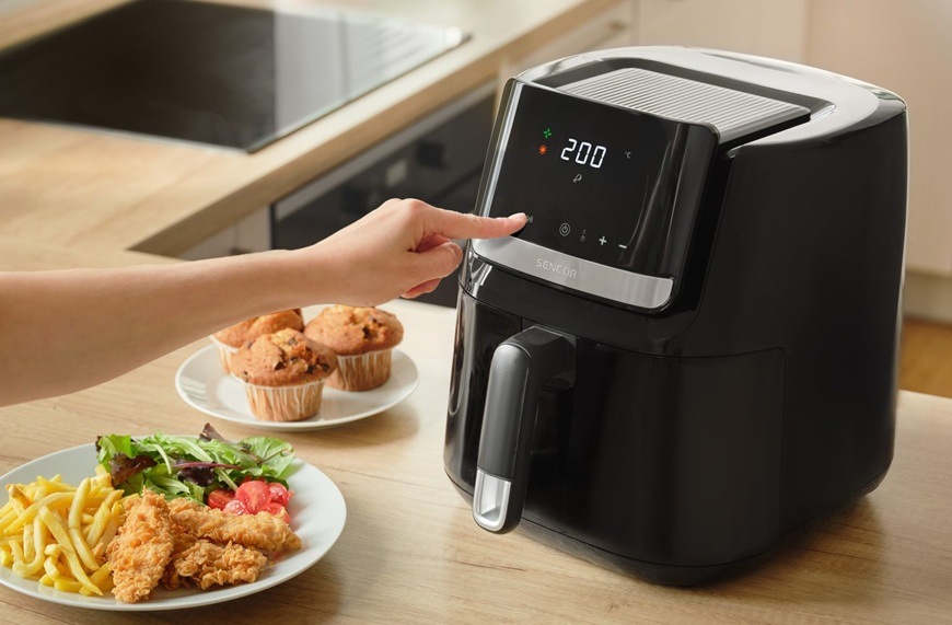 Fritéza teplovzdušná Sencor SFR 6550BK-EUE3 čierna Žena nastavuje na LED displeji pri teplovzdušnej fritéze značky Sencor časovač na 60 minút. Vedľa neho sa nachádzajú upečené muffiny a stripsy so zeleninou.
