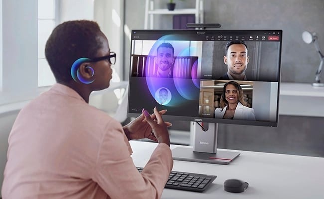 PC all in-one Lenovo ThinkCentre M90a Pro Gen 6 čierny/sivý Osoba so slúchadlami sedí pri stole pred počítačovým monitorom, na ktorom je vidieť niekoľko účastníkov videohovoru. Prostredie vyzerá ako moderné kancelárske prostredie s prirodzeným svetlom.