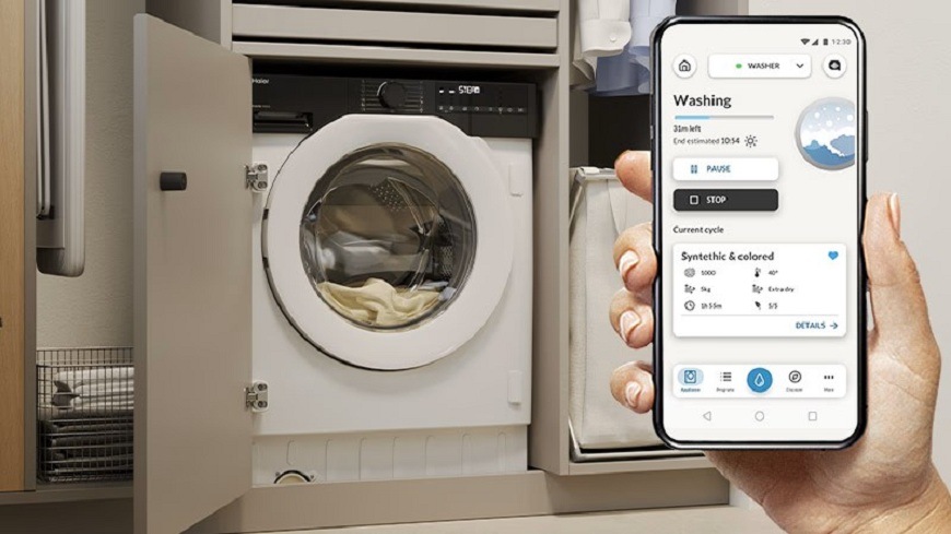 ruka drží telefón so zapnutou aplikáciou hon, v pozadí je práčka Candy BC4S68M6D8J-S s bielizňou