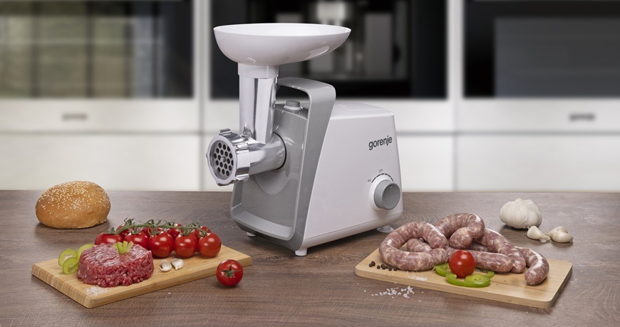 Mlynček na mäso Gorenje MG1601W je položený na stole a okolo neho sú rozmiestnené rôzne druhy pokrmov.