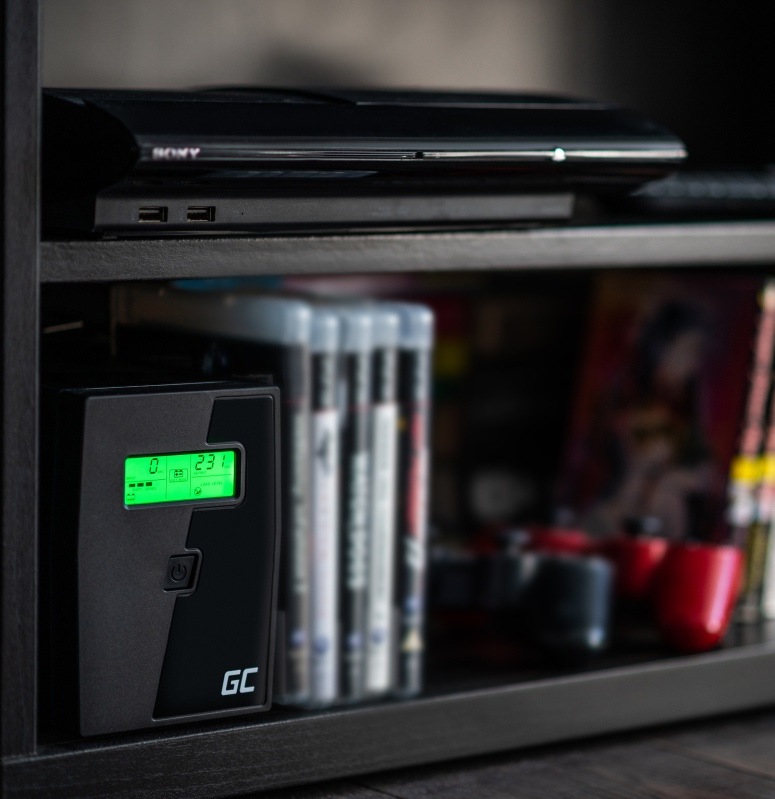 Záložný zdroj Green Cell Microsine 2000VA LCD čierny Na obrázku je záložný zdroj Green Cell Microsine umiestnený v polici domácej zostavy. Stojí vľavo dole na tmavej polici, má čierne hranaté telo a na čelnej strane svieti malý zelený displej s prevádzkovými údajmi. Pod displejom je tlačidlo zapnutia a dole logo GC. Nad ním je položené ďalšie elektronické zariadenie, okolo je vidieť obaly diskov a drobné predmety. Fotka ukazuje, že zdroj môže nenápadne fungovať aj v obývacej izbe alebo pri domácej technike.