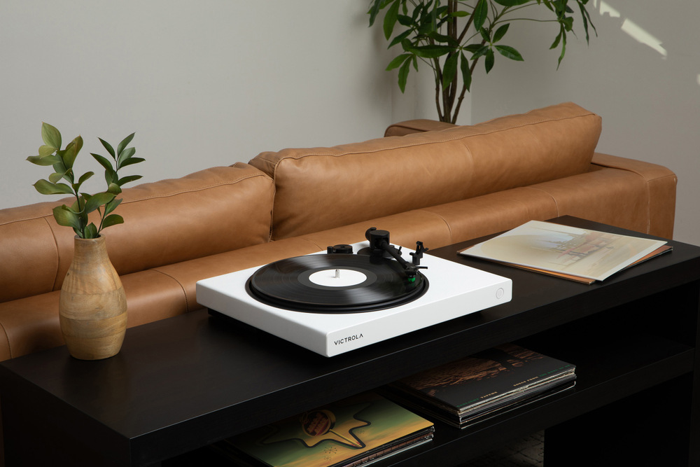 Biely gramofón Victrola VPT-1520 Wave umiestnený na stolíku v obývacej izbe, prehráva vinylovú dosku v modernom interiéri