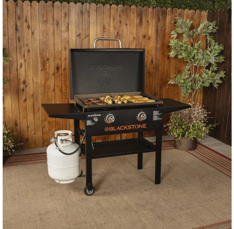 Gril záhradný plynový Blackstone 28" Griddle BS-2147 Blackstone 28" griddle v záhrade pri drevenom plote s grilovaným mäsom a propánovou fliaš.
