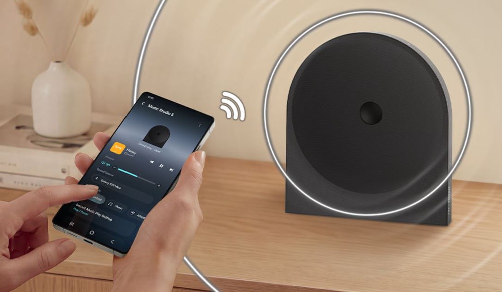 Reproduktor Samsung Music Studio 5 LS50H (1 ks) čierny ovládanie reproduktora Samsung HW-LS50H Studio 5 cez mobilnú aplikáciu v telefóne. Reproduktor je na drevenom stolíku, v pozadia je väza so suchou kvetinou.