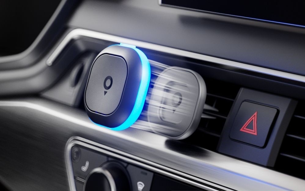 Cestovný asistent TomTom Tom pripevnený magnetickým držiakom k ventilačnej mriežke automobilu. Modro svietiaci LED krúžok signalizuje aktívne upozornenie a rozmazaný obrys znázorňuje ľahké sňatie zariadenia z držiaka.