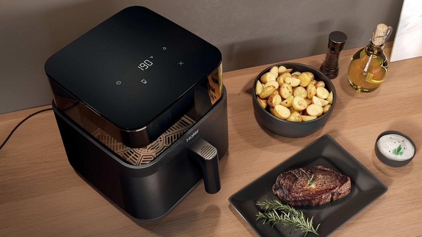 Fritéza teplovzdušná Haier I-Master Series 3 HAF3AFW 011 čierna Teplovzdušná fritéza Haier HAF3AFW 011 so zapnutým svetlom na drevenom stole, okolo nej sú zhotovené pokrmy a dochucovadlá