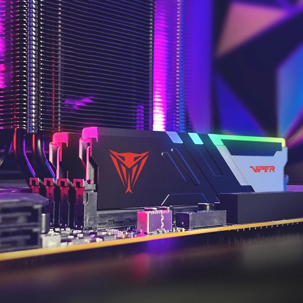 Pamäťový modul Patriot Viper Venom RGB 32GB (2x16GB) DDR5 6000MHz CL30 Na obrázku je pamäťový modul Patriot Viper Venom RGB osadený v základnej doske počítača. Modul má čierny chladič so strieborným detailom, červeným logom Viper a farebne svietiacou RGB lištou na hornej hrane. V pozadí je vidieť veľký chladič procesora s kovovými rebrami a celé okolie je nasvietené fialovým a ružovým RGB svetlom. Snímka ukazuje pamäť priamo v zostavenej počítačovej skrini.