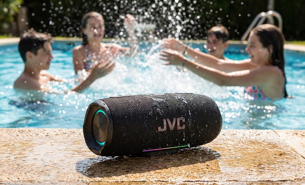 Vodeodolný prenosný Bluetooth reproduktor JVC XS-E425B pri bazéne so striekajúcou vodou a deťmi v pozadí