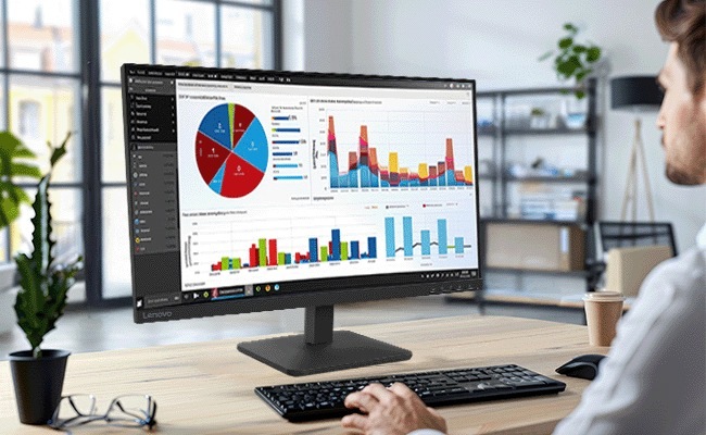 Monitor Lenovo L27-4e čierny Osoba v kancelárii si prezerá dátové grafy na širokouhlom monitore, zobrazujúce farebné grafy a koláčové grafy. Medzi predmety na stole patrí klávesnica, hrnček a okuliare.