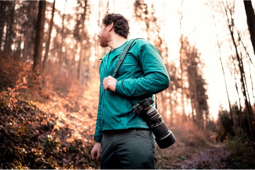 Muž v lese nesúci fotoaparát s objektívom Tamron 150–500 mm f/5–6.7