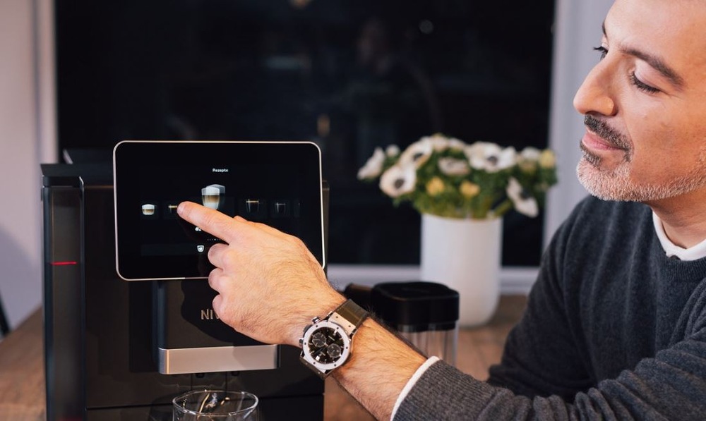 Starnúci muž so šedými fúzmi a hodinkami na ruke vyberá z menu na kávovare Nivona nápoj, ktorý si pripraví.