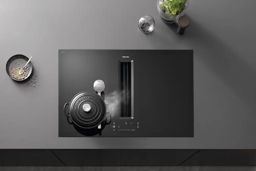 Na varnej doske Miele KMDA 7676-1 FL BlackPerfection stojí hrniec, para z hrnca je odsávaná integrovaným odsávačom, vedľa dosky leží miska jedlom