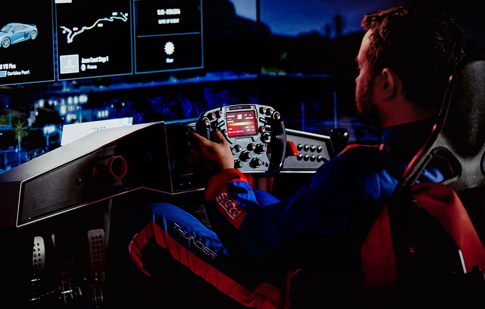 Základňa Moza Racing R25 Ultra Direct Drive hliník Osoba v závodnej kombinéze sedí v vodičskom simulátore a drží volant. Na niekoľkých obrazovkách sa zobrazujú závodné dáta a mapy tratí, čo vytvára sústredenú a pohlcujúcu atmosféru.