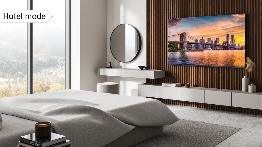 JVC TV v modernej, svetlom hotelovej izbe a veľkou posteľou. Na televízii je zobrazený západ slnka nad Brooklynským mostom v NYC. V ľavom hornom rohu je popis 'Hotel mode' zdôrazňujúci režim pre hotely