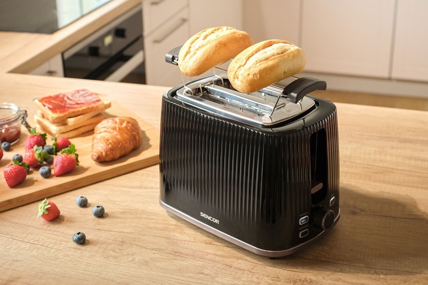 Hriankovač Sencor STS 7511BK čierny Na linke v modernej kuchyni leží hriankovač Sencor STS 7511BK. Vedľa neho je na doske položený croissant, džem, jahody a čučoriedky.