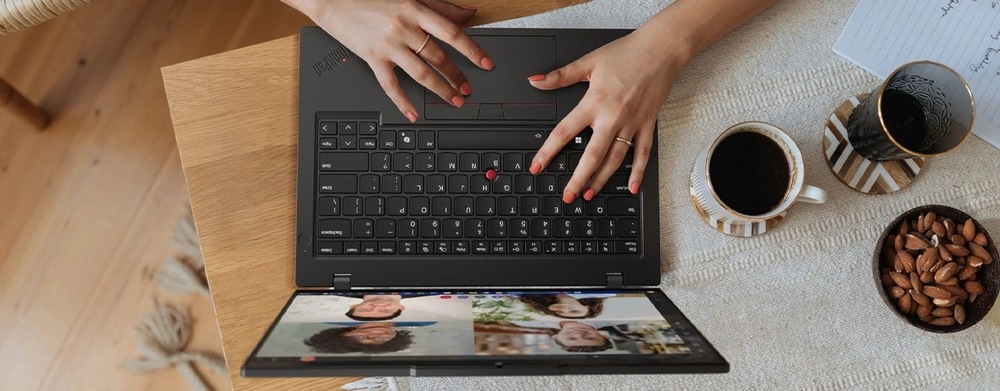 Notebook Lenovo ThinkPad L14 Gen 6 čierny Užívateľ píše na notebooku, na ktorom je vidieť videohovor so štyrmi ľuďmi. Vedľa neho stojí šálka čiernej kávy, mandle a zápisník, ktoré vytvárajú útulnú a produktívnu atmosféru.