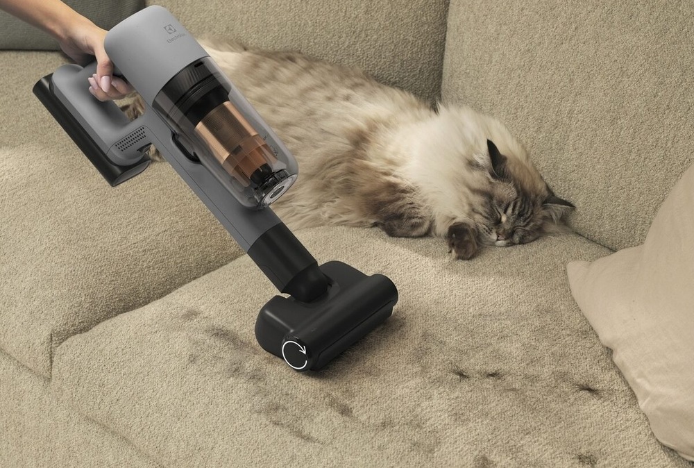 Tyčový vysávač Electrolux 700 Animal EP72AB21UG sivý Mačka leží na pohovke plnej chlpov. Vedľa nej žena s ručnou jednotkou vysávača Electrolux a hubicou PetPro+ vysáva chlpy.