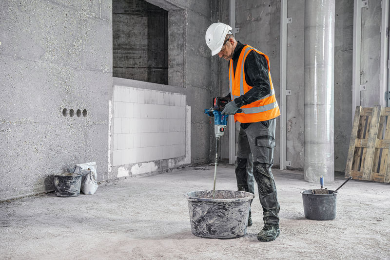 Pohľad na muža na stavbe, ktorý v rukách drží miešadlo Bosch Professional GRW 18V-160 a mieša vo vedre cement.