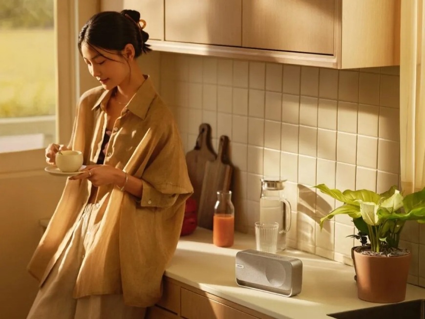 Prenosný reproduktor Bose Soundlink HOME sivý Žena so šálkou kávy v ruke sa opiera o kuchynskú linku, na ktorej je reproduktor Bose SoundLink Home