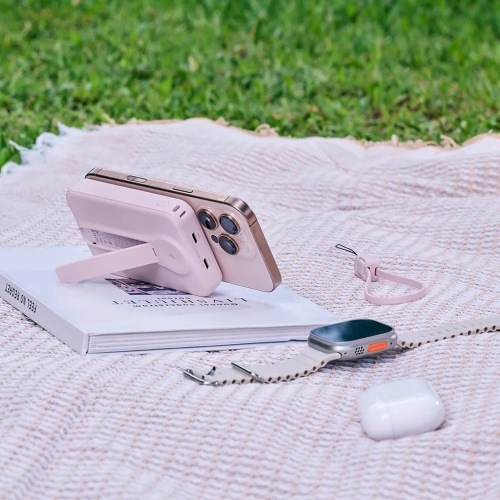 Piknik na trávniku: powerbanka Adam Elements v ružovej drží iPhone na stojane vedľa Apple Watch a AirPods.