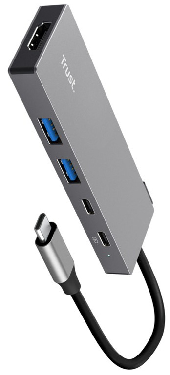 Na obrázku je strieborný USB-C hub Trust DALYX 5v1 zachytený šikmo spredu. Má úzke kovové telo s jednoduchým hranatým tvarom a z jedného konca z neho vedie krátky kábel zakončený USB-C konektorom. Na boku sú dva modro označené USB porty, na čele HDMI konektor a na ďalšej strane dva menšie USB-C porty.