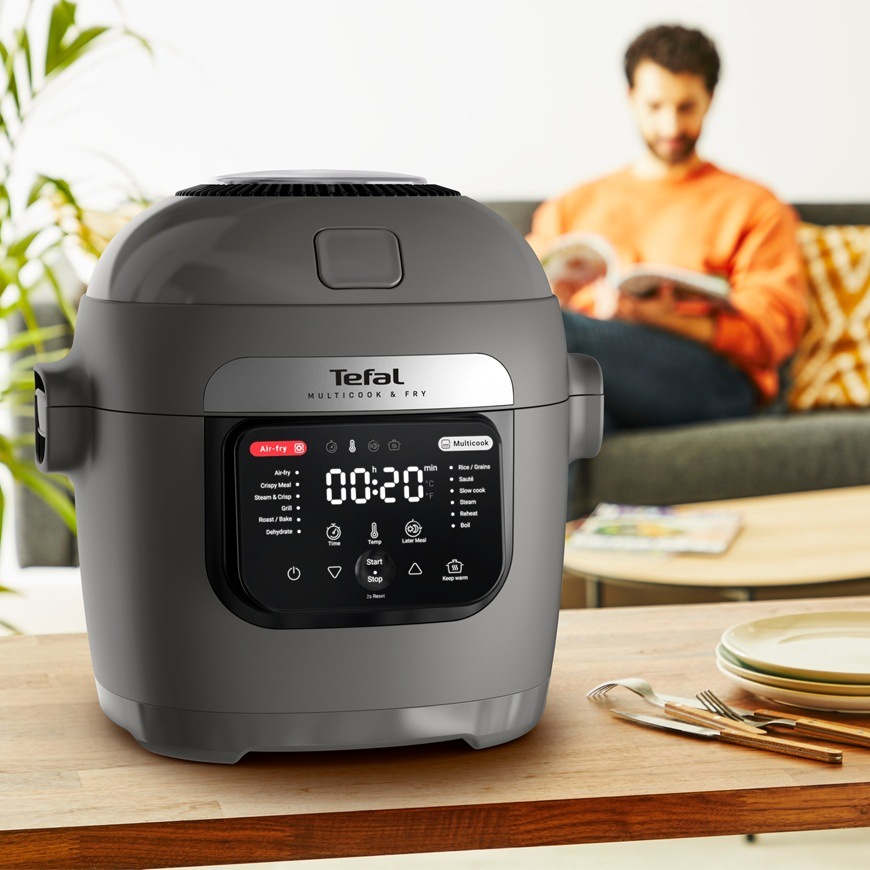 Multifunkčný hrniec Tefal MY731BF0 Multicook & Fry sivý Multifunkčný hrniec Tefal MY731BF0 s digitálnym displejom stojí na stole v obývacej izbe, zatiaľ čo muž v pozadí číta časopis na pohovke.