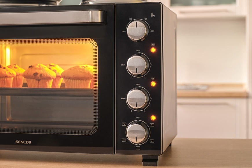 Detail mini rúry Sencor SEO 3500BK s osvetleným vnútrom pri pečení muffinov, doplnený o otočné ovládače na nastavenie teploty, časovača a režimov pečenia.