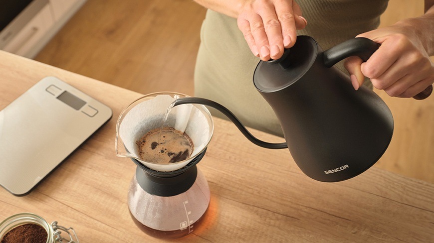 Rýchlovarná kanvica Sencor SWK 2828BK, 0,6 l čierna Žena si pripravuje pomocou rýchlovarnej kanvice Sencor SWK 2828BK prekvapkávanú kávu. Na linke stojí aj kuchynská váha.