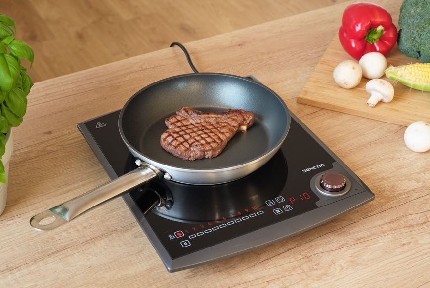 Varič indukčný Sencor SCP 3414GY čierny/sivý Indukčný varič Sencor SCP 3414GY s dotykovým ovládaním a otočným regulátorom pripravuje steak na panvici na svetlej drevenej kuchynskej linke zatiaľ čo v pozadí sú čerstvé suroviny ako paprika brokolice kukurice a šampiňóny na doske.