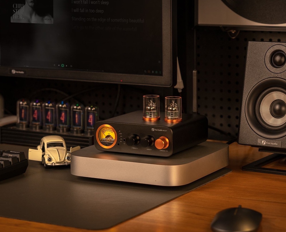 Zosilňovač Fosi Audio MC331 čierny Na obrázku je zosilňovač Fosi Audio MC331 umiestnený v pracovni alebo domácom posluchovom kútiku. Stojí na stole na striebornom podstavci, má čierne telo, dve elektrónky na hornej strane a výrazný oranžovo podsvietený VU merač aj hlavný ovládač na prednom paneli. Vpravo vedľa neho je reproduktor, vľavo monitor a ďalšia drobná elektronika, takže celá scéna pôsobí ako štýlovo zariadené miesto na počúvanie hudby. Hore je anglický reklamný text, ktorý zdôrazňuje kompaktné rozmery a schopnosť zapadnúť do rôznych audio zostáv.