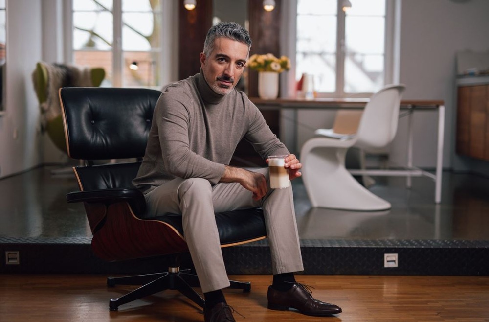 Starší muž so šedými vlasmi a fúzmi sedí na koženom kresle, v ruke má pohár s latté. V pozadí je biela stolička, stôl a dve okná.