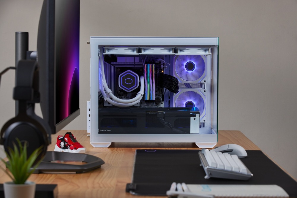 Elegantná biela počítačová skriňa s osadenými komponentmi, svietiacimi fialovými LED ventilátormi a káblami na drevenom stole. Monitor, klávesnica a slúchadlá sú blízko a vytvárajú modernú zostavu.