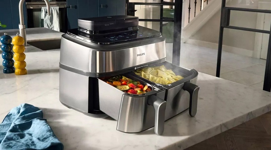 Fritéza teplovzdušná Philips Airfryer Dual Basket 5000 series 9 l, NA555/00 strieborná Teplovzdušná fritéza Philips s dvoma vysunutými košmi umiestnená na kuchynskej linke, v jednom koši je pripravovaná zelenina av druhom zemiaky, z ktorých stúpa para.